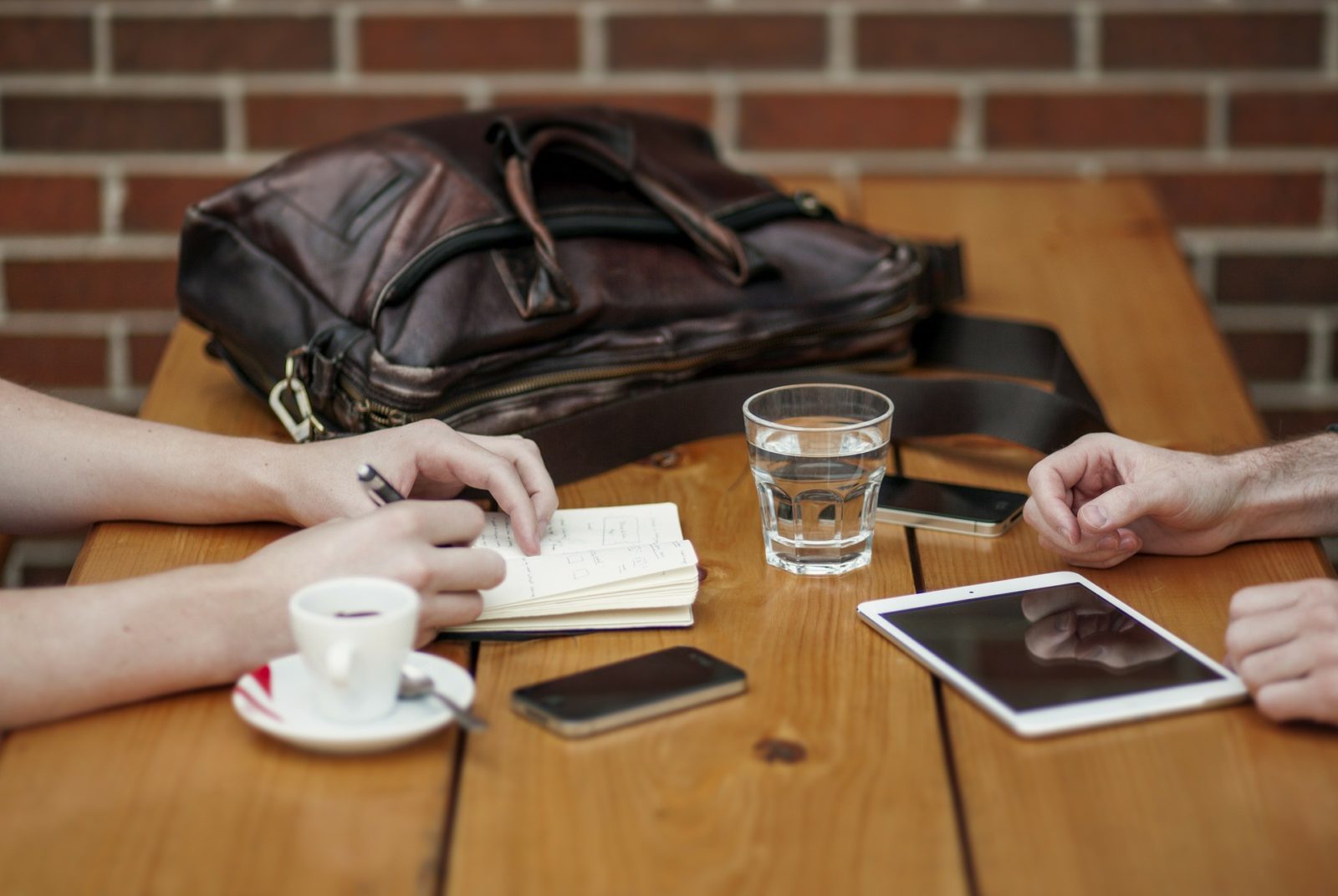 Two people sit opposite each other in a sales conversation at a table with drinks, mobile devices, a bag and a notebook arrayed on top.